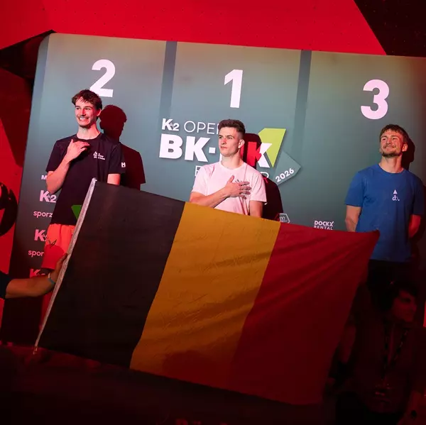 Drie mannelijke atleten staan op een podium met bordjes voor de eerste, tweede en derde plaats. De atleet in het midden legt zijn hand op zijn hart, terwijl een andere atleet een grote Belgische vlag voor zich houdt.
